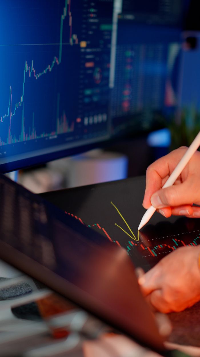 about-03 Person using a stylus on a tablet for stock market analysis, with charts on a monitor.